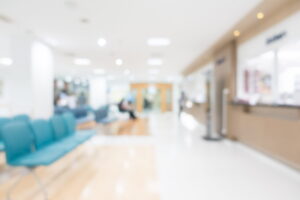 Blurred view of a bright, modern medical clinic waiting room with teal chairs in the foreground and a reception desk on the right.
