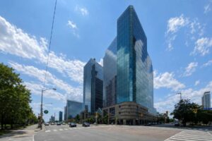 Two modern blue glass office towers stand at a wide city intersection under a bright, partly cloudy sky with light traffic on the road.