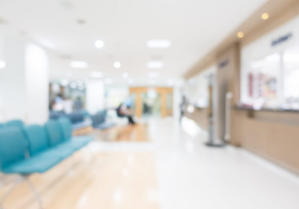 Blurred view of a bright, modern medical clinic waiting room with teal chairs in the foreground and a reception desk on the right.