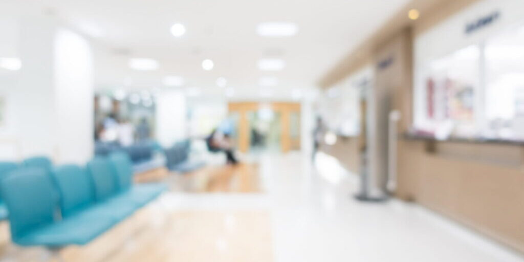 Blurred view of a bright, modern medical clinic waiting room with teal chairs in the foreground and a reception desk on the right.