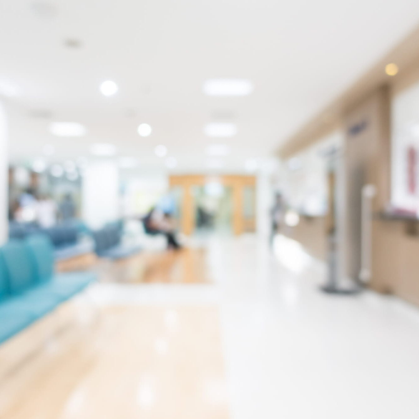 Blurred view of a bright, modern medical clinic waiting room with teal chairs in the foreground and a reception desk on the right.