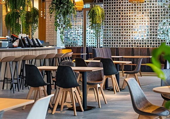 Modern cafe and restaurant interior featuring light-wood tables, black shell-style dining chairs, and gray upholstered seating. A long service bar with a light wood base and matching bar stools with black legs sits to the left. The stylish space is decorated with lush hanging green plants, warm pendant lighting, a textured privacy screen, and spotless tile flooring, representing a clean hospitality environment maintained by professional restaurant drain cleaning and sanitization.