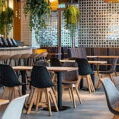 Modern cafe and restaurant interior featuring light-wood tables, black shell-style dining chairs, and gray upholstered seating. A long service bar with a light wood base and matching bar stools with black legs sits to the left. The stylish space is decorated with lush hanging green plants, warm pendant lighting, a textured privacy screen, and spotless tile flooring, representing a clean hospitality environment maintained by professional restaurant drain cleaning and sanitization.