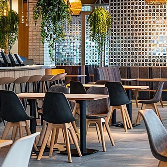Modern cafe and restaurant interior featuring light-wood tables, black shell-style dining chairs, and gray upholstered seating. A long service bar with a light wood base and matching bar stools with black legs sits to the left. The stylish space is decorated with lush hanging green plants, warm pendant lighting, a textured privacy screen, and spotless tile flooring, representing a clean hospitality environment maintained by professional restaurant drain cleaning and sanitization.